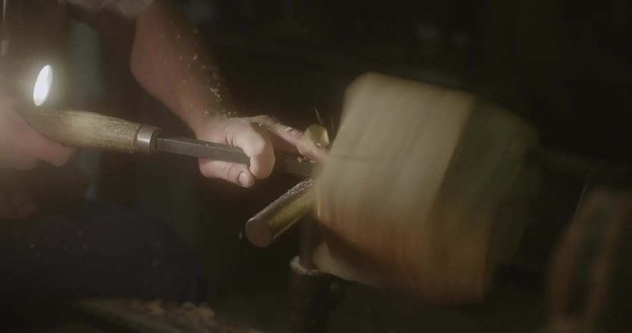 A woodworker holds a chisel as they carve at a piece of wood that is spinning in a lathe