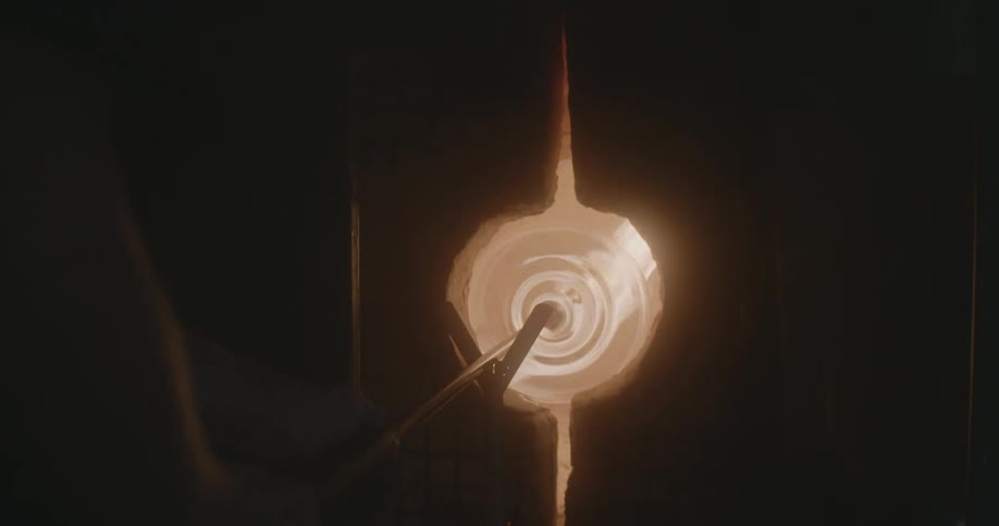 A glassmaker holds a pole with hot glass in the oven propped up to hold it steady