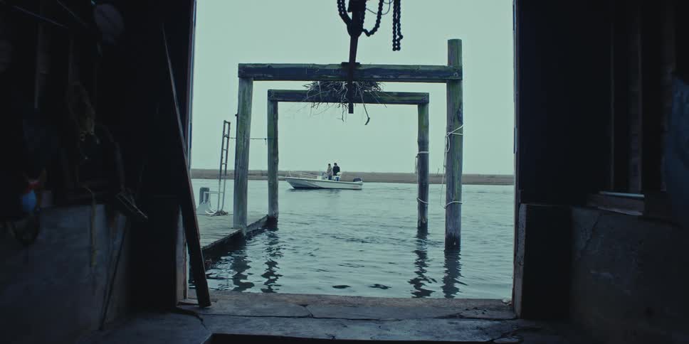 Two people stand in the back of a boat as it sails through a lake near a pier