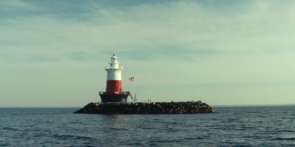 An american flag flies on a barge parked in front of a lighthouse