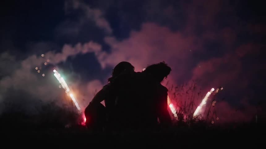 The silhouette of a couple sitting cuddled on the grass watching fireworks at night