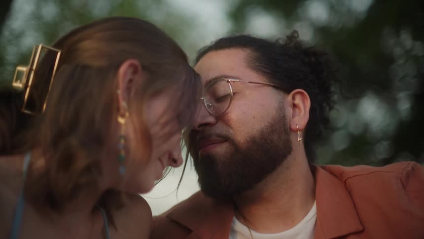 A man and a woman are sitting outside together looking into each others eyes and he kisses her on the forehead