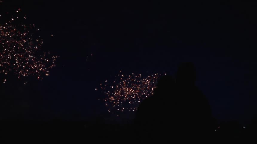 Silhouettes of a couple sitting together in the grass watching bright and colorful fireworks