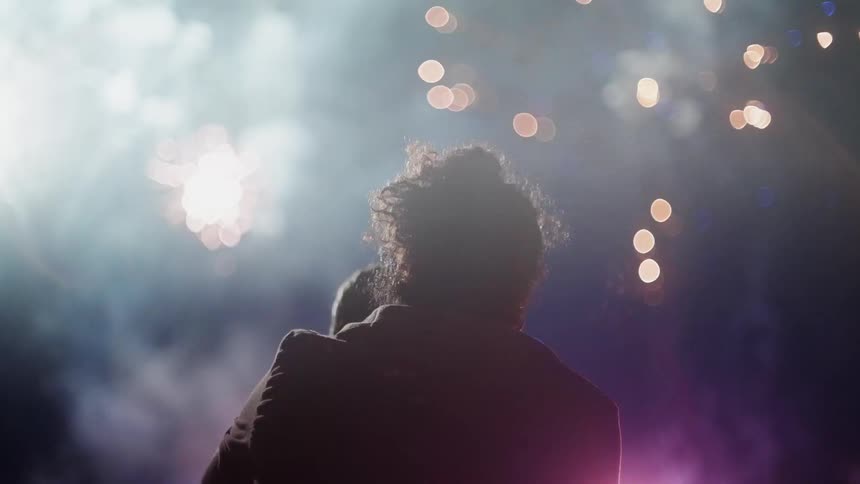 A man holds a woman in front of him as they watch fireworks light up the night sky