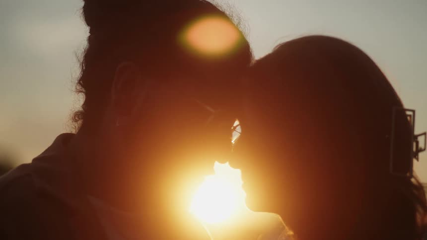 A man and woman stand with their noses together as the sun sets behind them