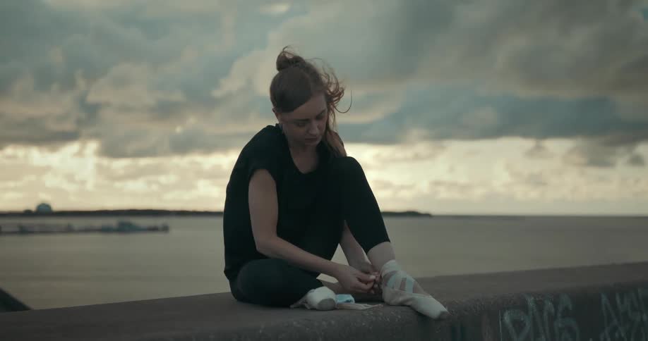 A woman sitting on a wall at the beach putting on ballet shoes