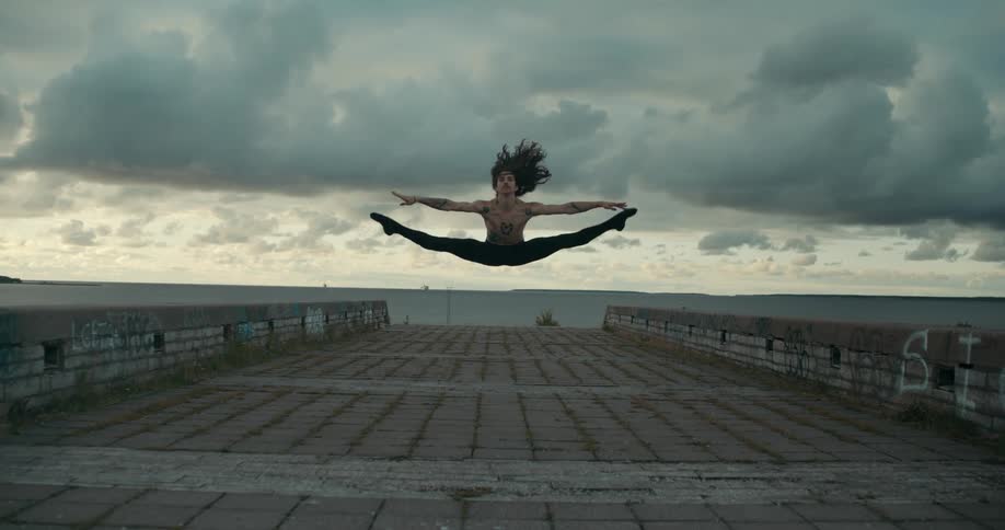 A man dancing on a pier jumps in the air and touches his toes as they go to the side of his body