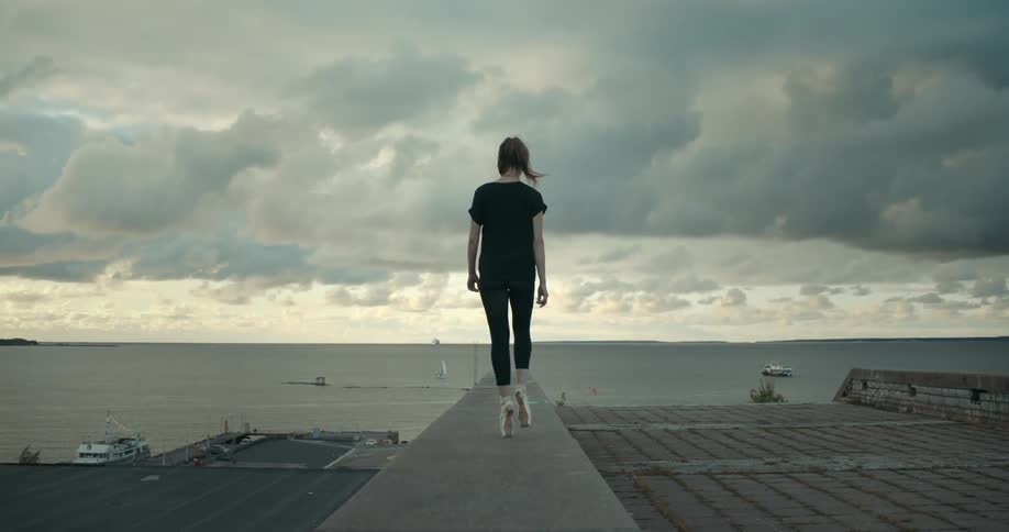 A ballet dancer walks along a ledge on a pier overlooking the sea