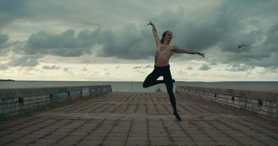 A man is standing on a bridge near the waterfront and he leaps into the air striking a dance pose before he lands as a seagull flies under a cloudy sky