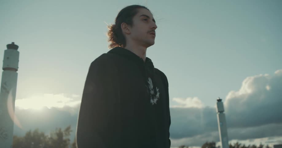 A man is standing outside at a park and he is moving his upper body as the sun shines though the clouds and the wind blows his hair
