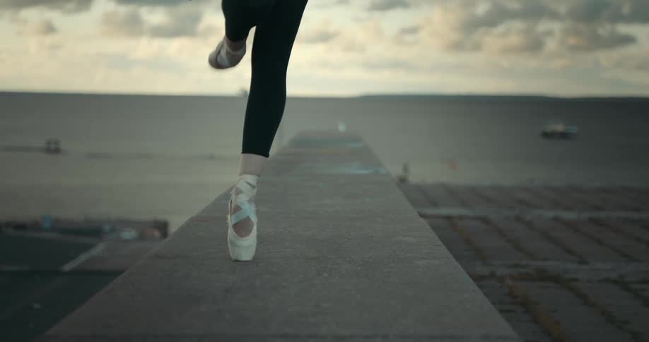 A woman wearing ballet pointe shoes walks on the top of a stone wall with the ocean and boats behind her