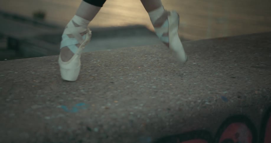 A dancer is walking across the top of a concrete wall on her toes in her ballet shoes