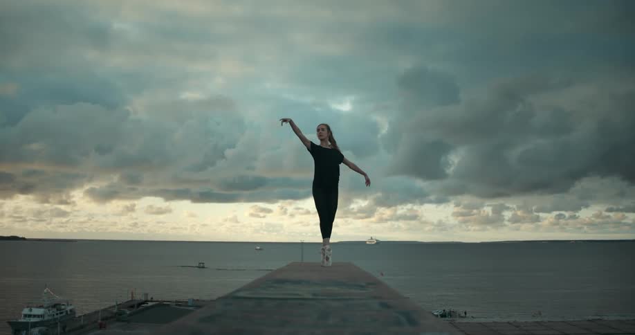 A woman is on a pier dancing on her toes under a cloudy sky as a seagull flies overhead