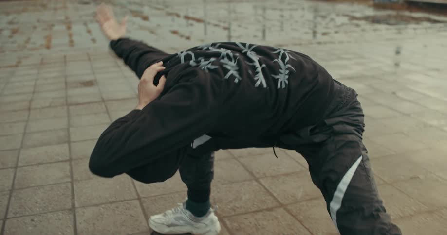 Person wearing hoodie moves through stretches in empty overgrown lot filled with pavers