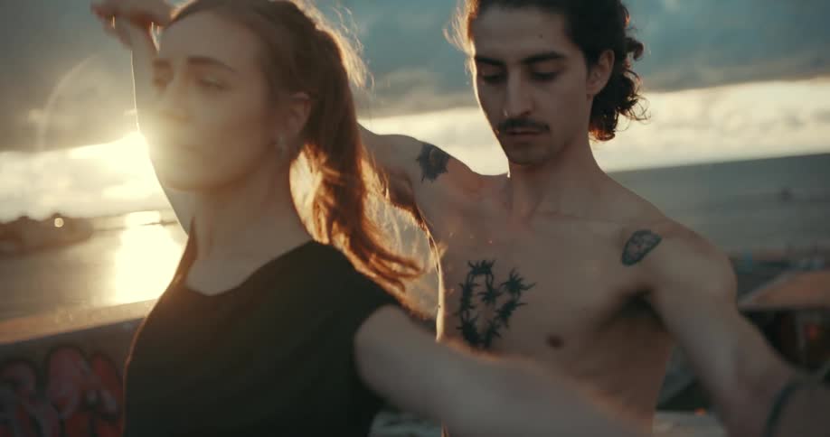 A man and a woman are dancing together on a bridge overlooking the water as the sun shines through a cloudy sky