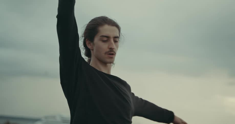 A man is standing along the waterfront practicing dance moves as seagulls fly overhead and a ship is in the water in the distance