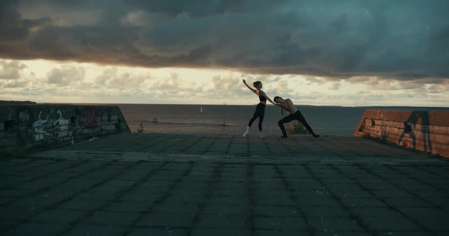 Sunlight casts the shadows of dancers dancing in a graffiti dotted paved area against the backdrop of the ocean