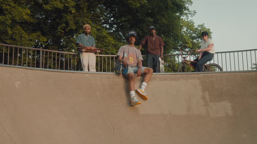 A teenage boy sits on a ledge holding his skateboard while two other teens stand behind him holding skateboards next to a girl on a bike