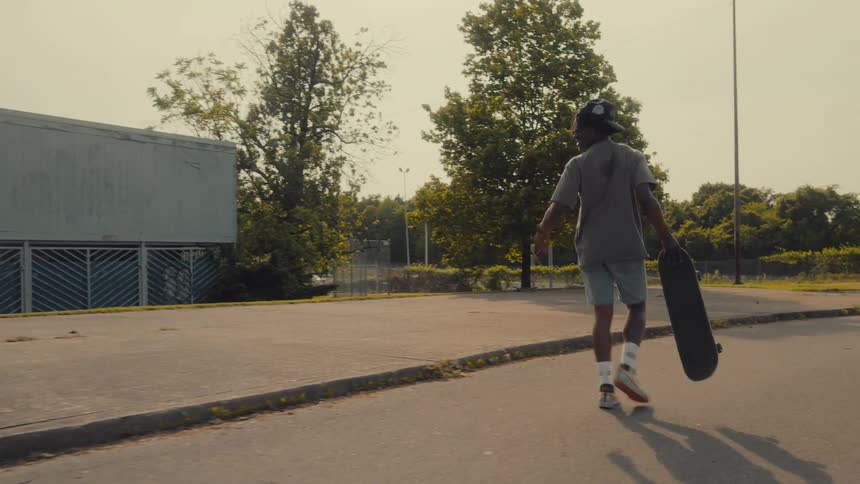 Man with cap purple shirt shorts white socks and sneakers carries skateboard on the street