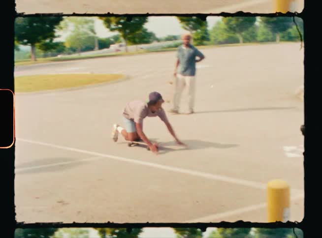 Vintage footage of a man holding a skateboard laughs as he watches his friend kneeling on a skateboarding and then falling off