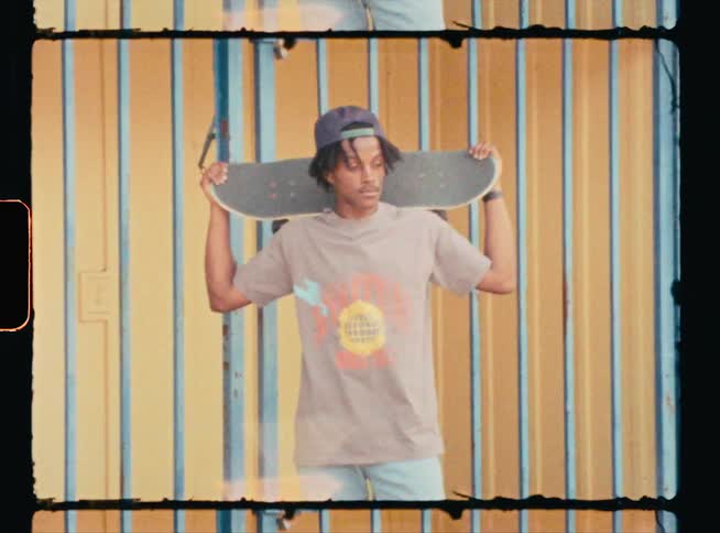 A young stands by a building holding a skateboard behind his head
