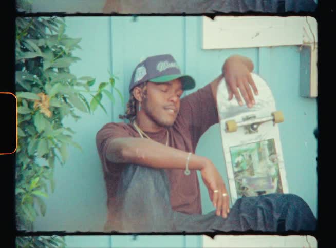 A man sitting outdoors with his back against a wall gestures at the camera while resting his elbow on a skateboard
