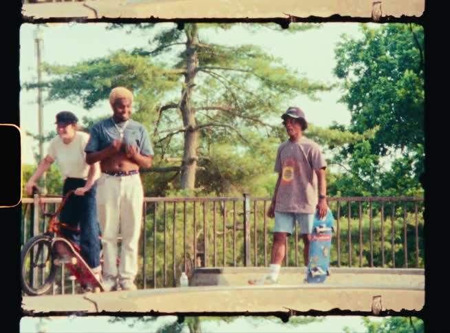Two teenage boys holding skateboards at a park talk to another boy as he wipes his face with his shirt as a girl on a bike laughs as she goes past