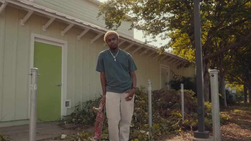 A young male poses while leaning on his skateboard outside of a building