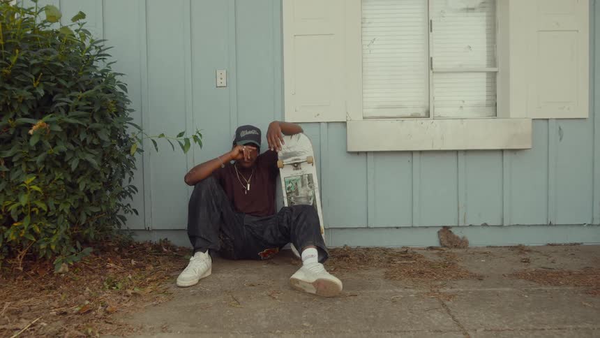 A man is sitting by a house with his skateboard