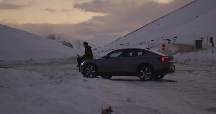 Man sits on the hood of his car with a laptop near the snowy mountains in the winter