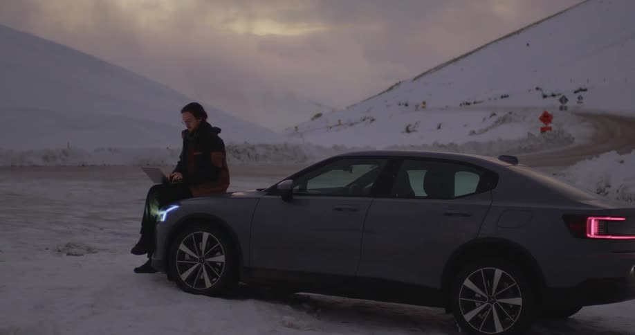 A man types on a laptop while sitting on the hood of a car parked beside a road on a snowy mountain
