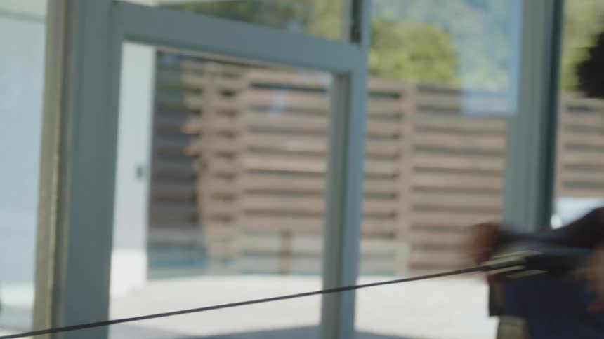 A man works out on a rowing machine at home near large windows looking out on a patio