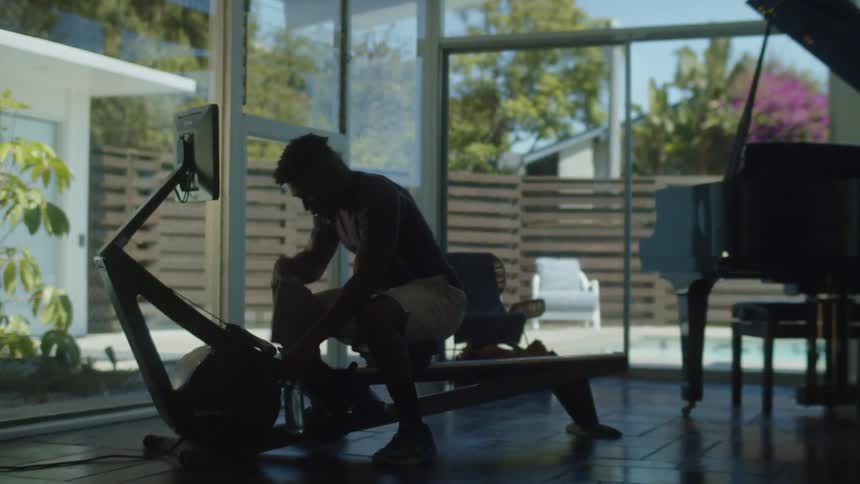 A man walks over to and sits down on a rowing machine at home