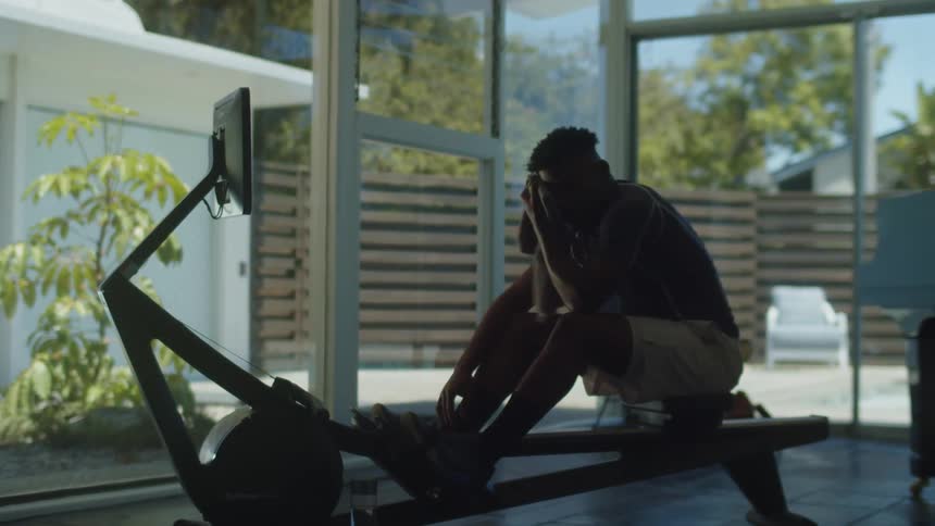 A man at home wipes his face as he sits on a rowing machine