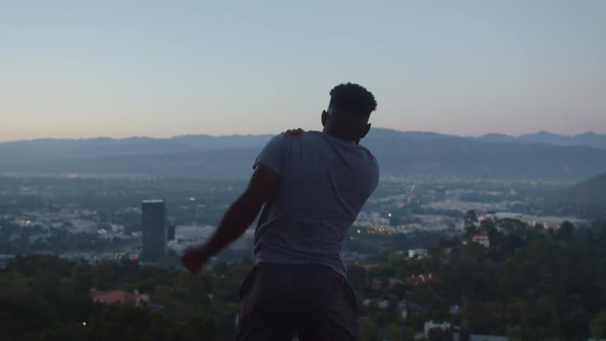 A man stretches his shoulder while standing on a hill overlooking a city during the day