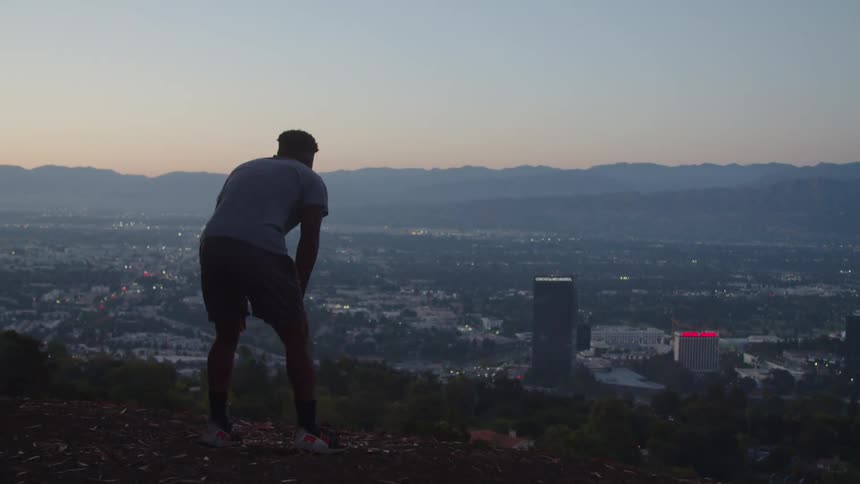 A man runs to the side of a hill overlooking a city below and there are mountains on the horizon