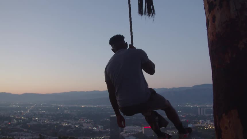 A man sitting on a rope swing swinging out over the city valley below