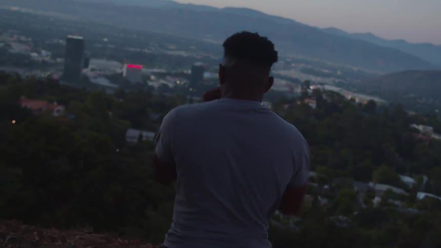 A man is moving his arms and exercising as he overlooks the city buildings below and the mountains on the horizon