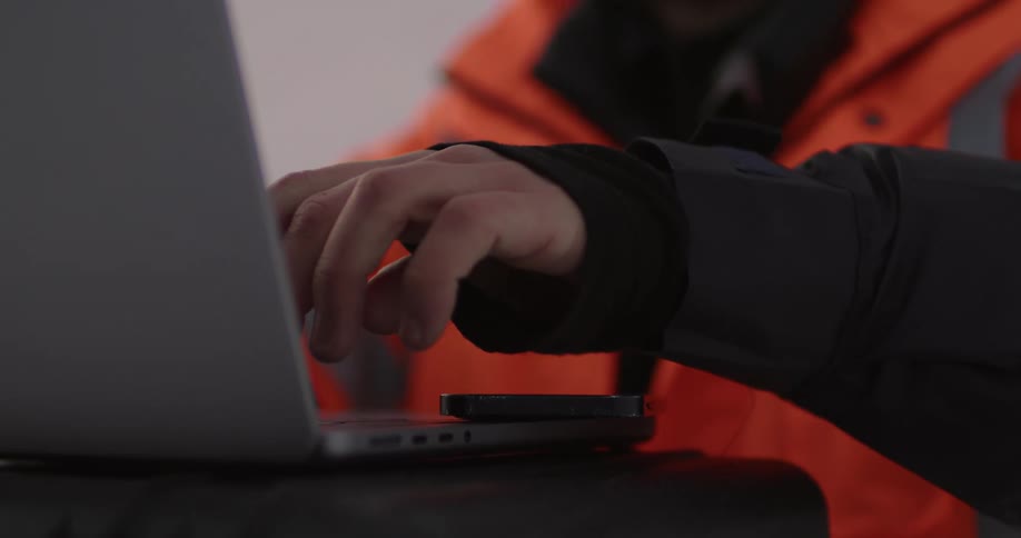 A man wearing a bright orange jacket is standing outside typing on a laptop computer and he rubs his hands together
