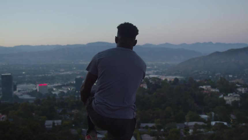 A man stretches his legs and knees while standing on a hill overlooking a city