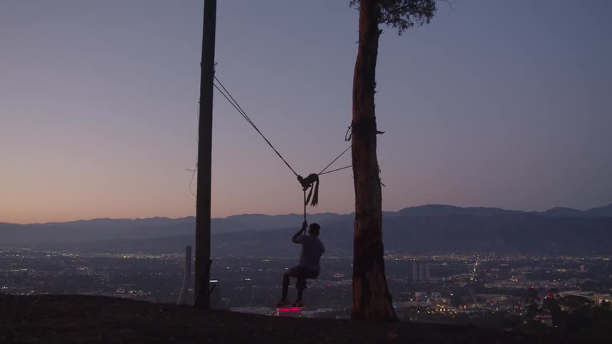 A man hangs from a zip line hung between trees overlooking a neighborhood