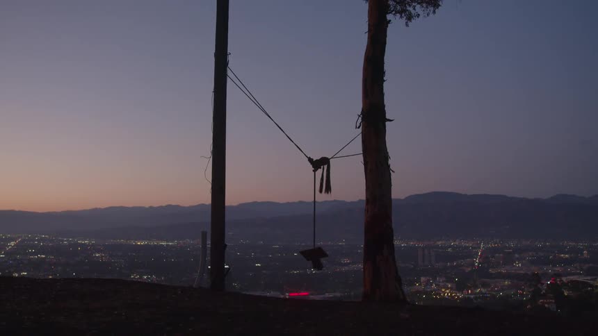 A swing tied between trees moves in the breeze as it overlooks a city below and mountains on the horizon