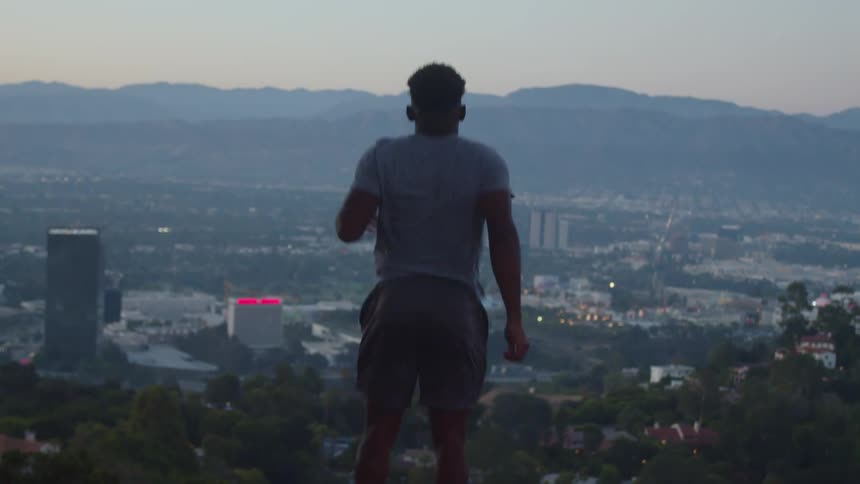 A man is jumping and exercising as he overlooks the city buildings below and the mountains on the horizon