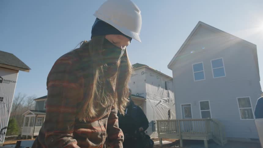 A female construction work is standing over a piece of wood and writing on it outside in front of a housing development they are building
