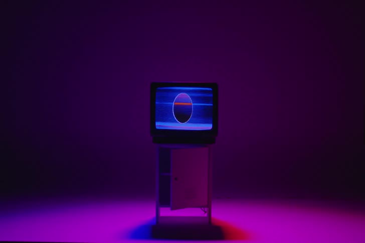 An oval shape in pink and blue static move on a television screen sitting on a floor illuminated with a magenta colored light