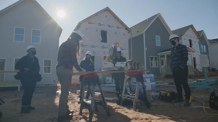 Construction workers stand around power tools sitting on boards propped up by sawhorses in dirt yards of houses under construction