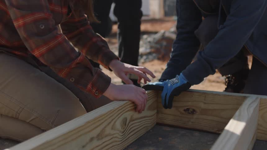 Two women kneel at a construction site and hold pieces of wood together as they discuss it and a man stands above them watching