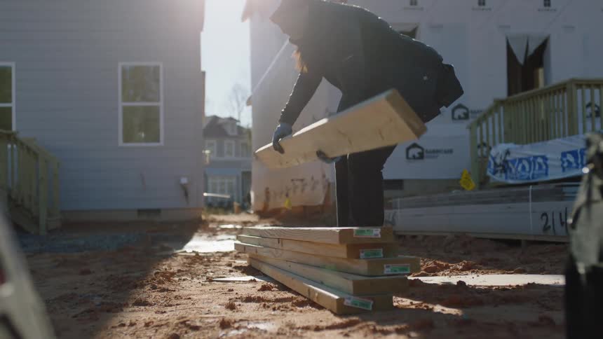 A construction worker carries plywood and stacks it on the ground