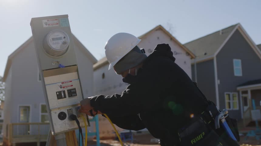 A construction worker plugs an electrical cord into an outlet and meter outside of a construction site in a neighborhood development