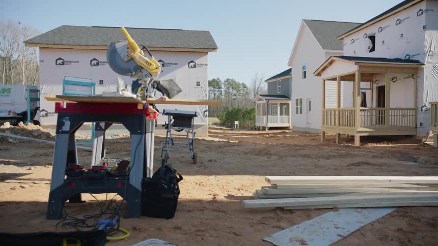A female construction worker picks up a wooden board and carries it across the construction site to a table with a saw